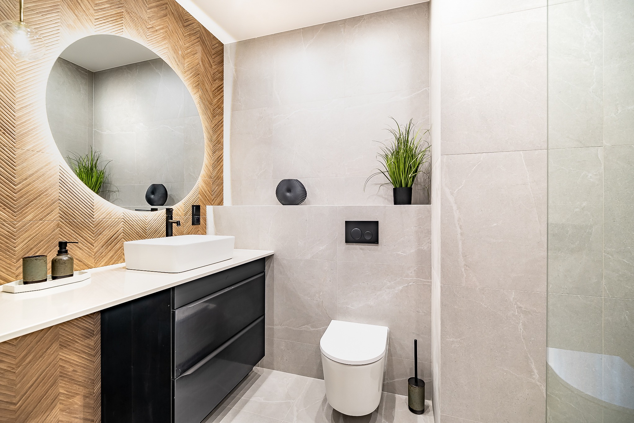 Nowoczesna łazienka z drewnianą mozaiką, okrągłym lustrem z podświetleniem LED, białą umywalką nablatową i szarą armaturą. Minimalistyczny design.