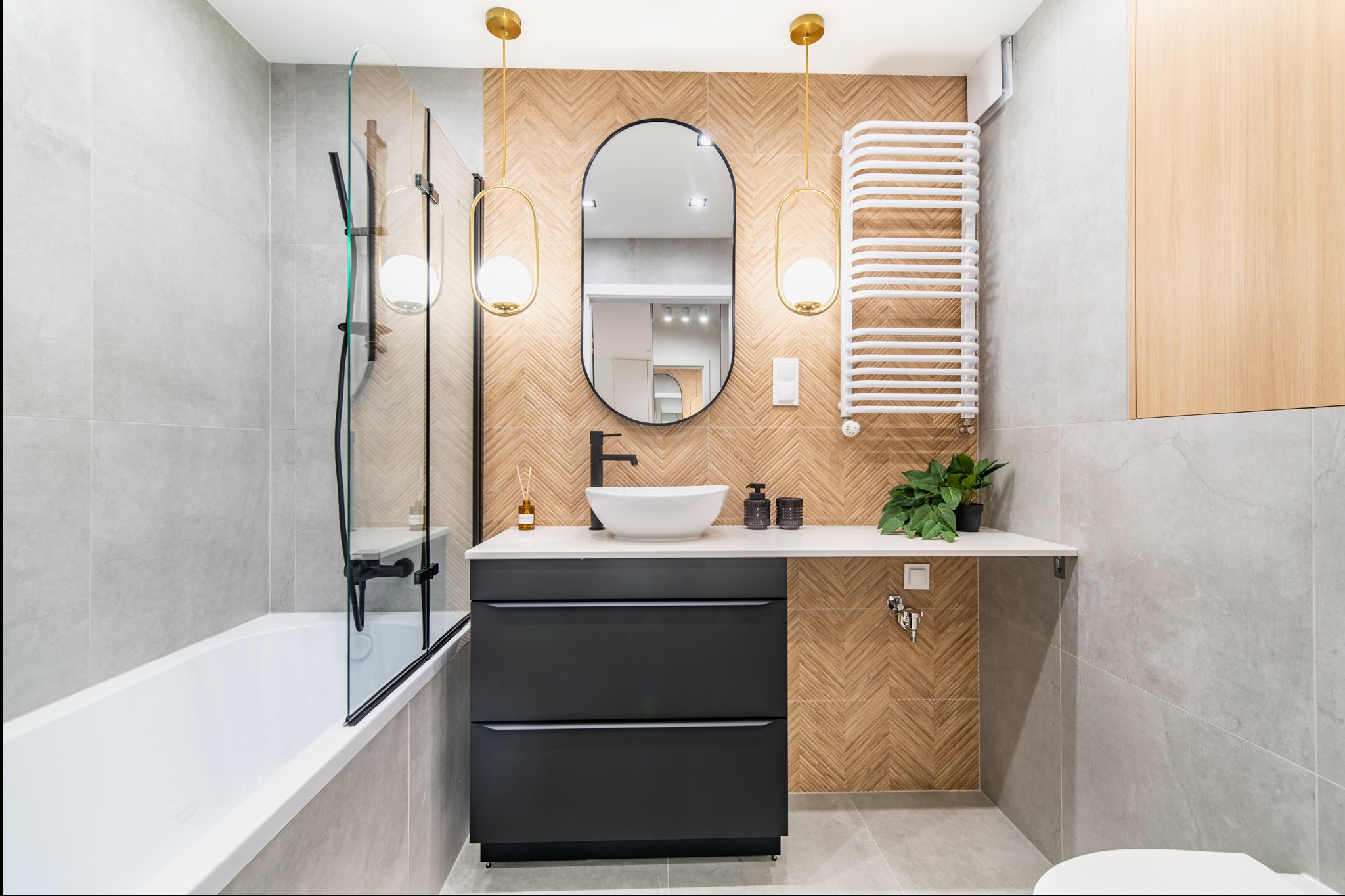 Nowoczesna łazienka z owalnym lustrem, drewnianą mozaiką na ścianie i czarną szafką pod umywalką. Minimalistyczny design, szare płytki i złote lampy.