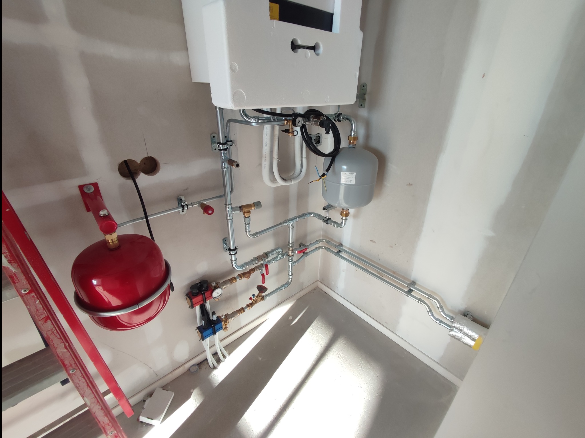 Złożona instalacja hydrauliczna z czerwonym zbiornikiem i białym piecem, rury prowadzone wzdłuż ściany, widoczne elementy montażowe i drabina. Szare ściany w tle.