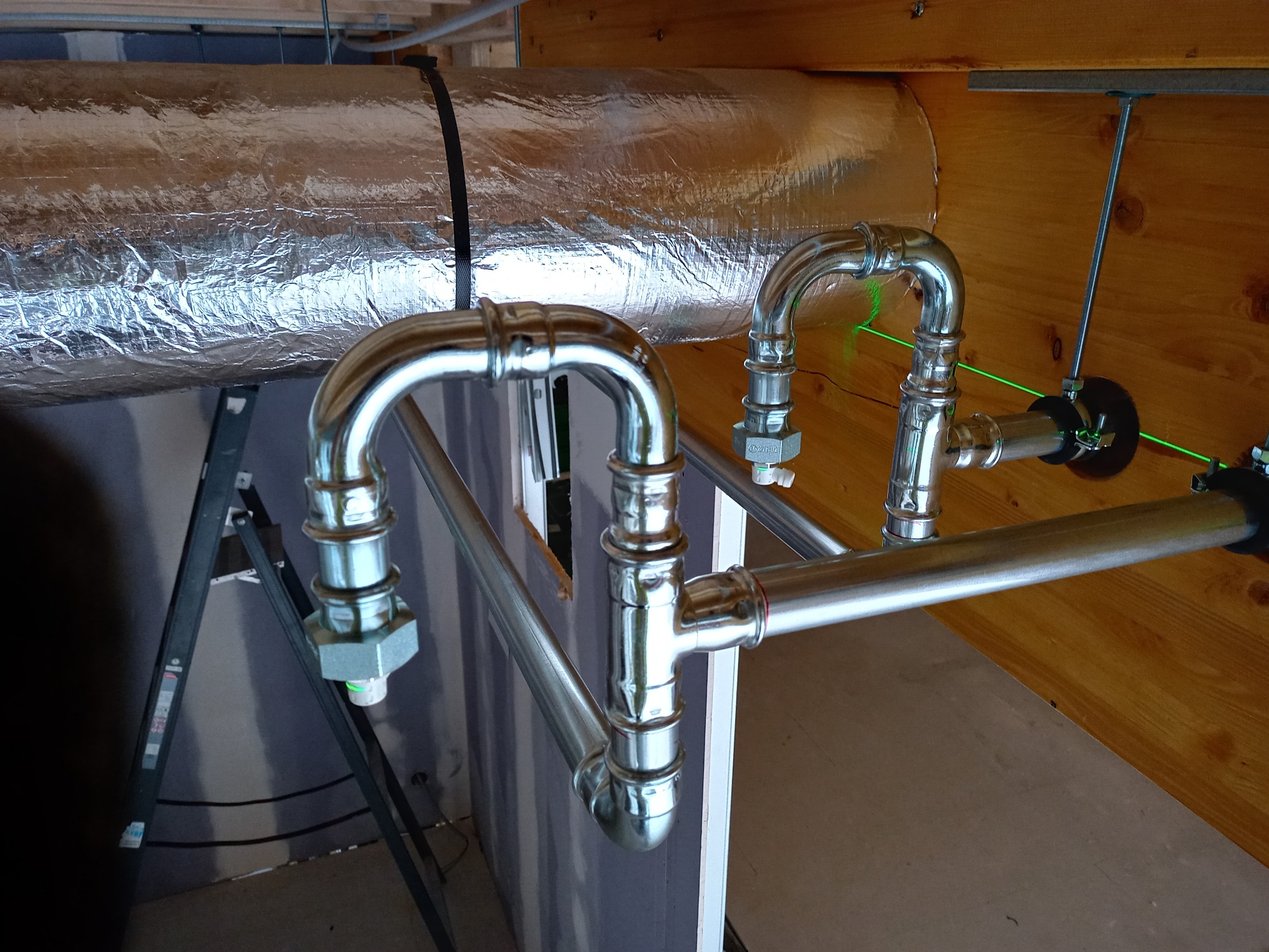 Błyszczące, chromowane rury instalacji wodnej, z zielonym laserem poziomującym, zamontowane pod drewnianym stropem. Widoczna izolacja termiczna rury wentylacyjnej.