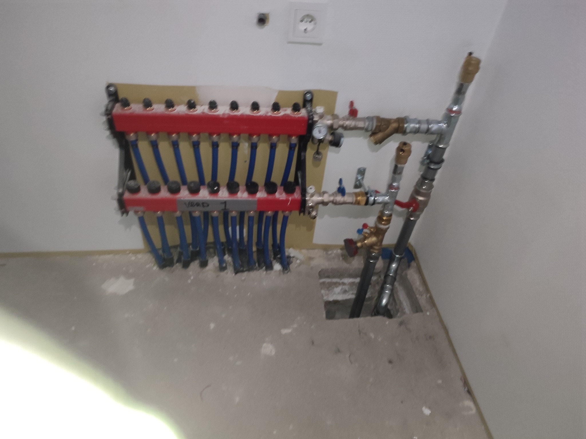 Rozdzielacz ogrzewania podłogowego z niebieskimi rurami, zamontowany na ścianie w surowym wnętrzu. Widoczne przyłącza i otwór w podłodze na rury.