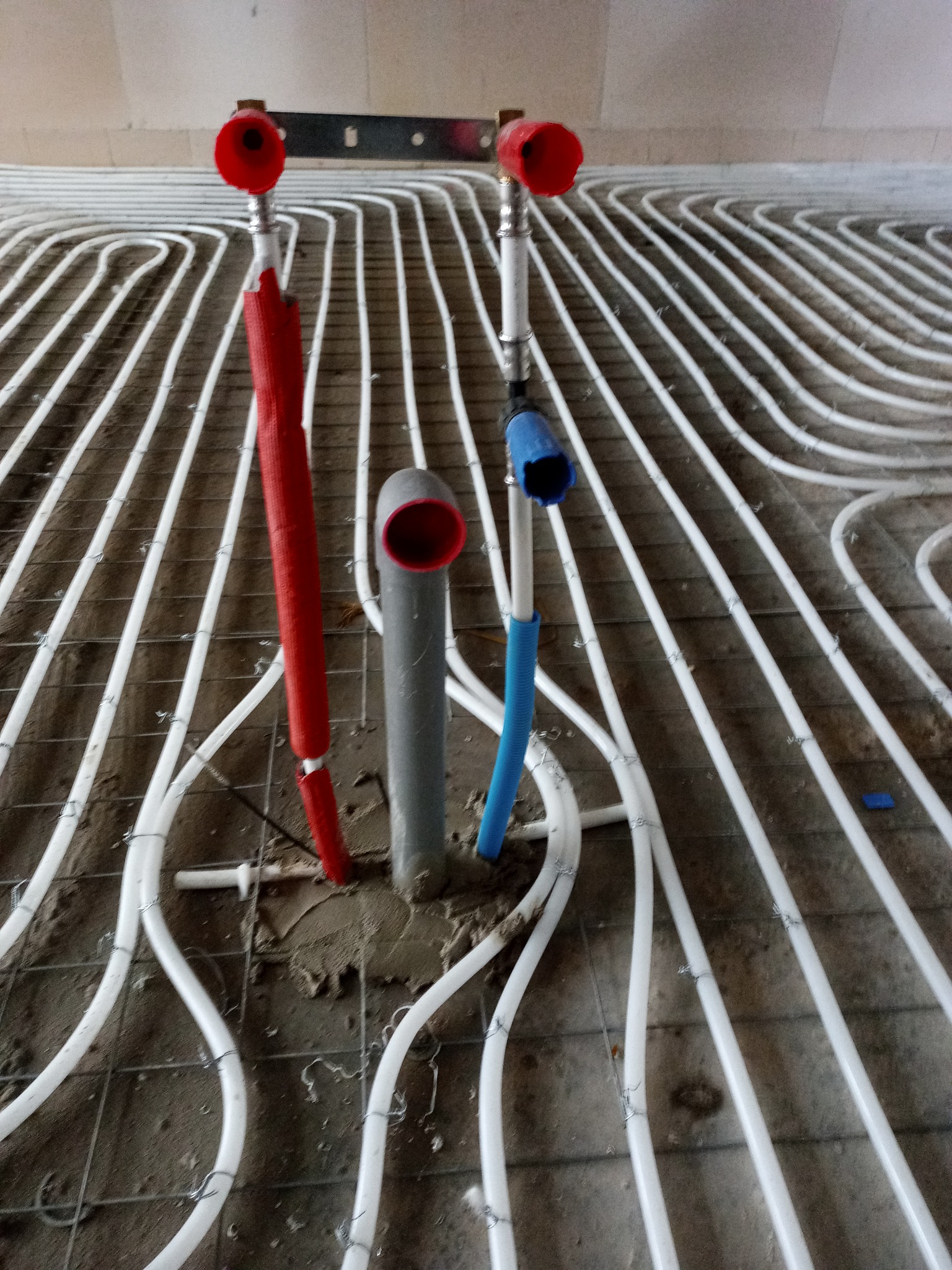 Białe rury ogrzewania podłogowego ułożone na siatce, z wystającymi rurami pionowymi w kolorach: czerwonym, szarym i niebieskim, przed wylaniem posadzki.