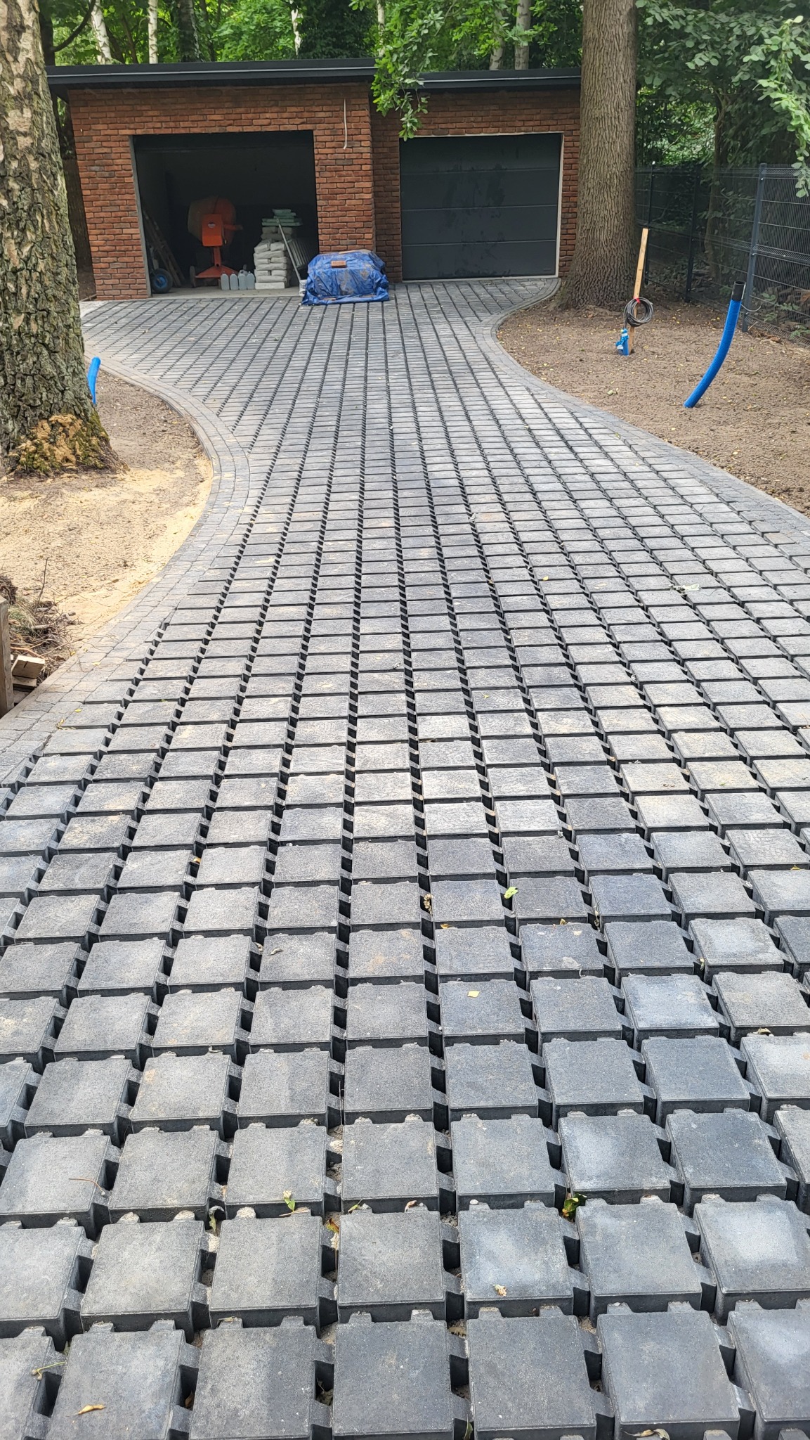 Ścieżka z ażurowych, szarych płyt betonowych prowadząca do garażu. Widoczne drzewa i elementy otoczenia. Charakterystyczny wzór płyt z otworami.