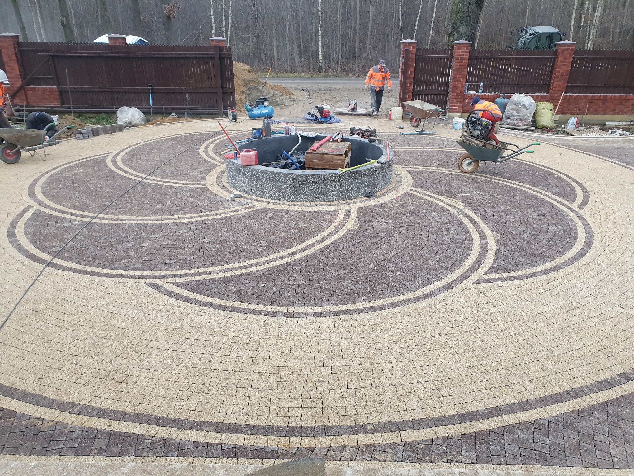 Okrągły podjazd z kostki brukowej ułożonej we wzór spirali, otoczony betonowym obrzeżem. W tle widoczne narzędzia i pracownicy podczas realizacji.