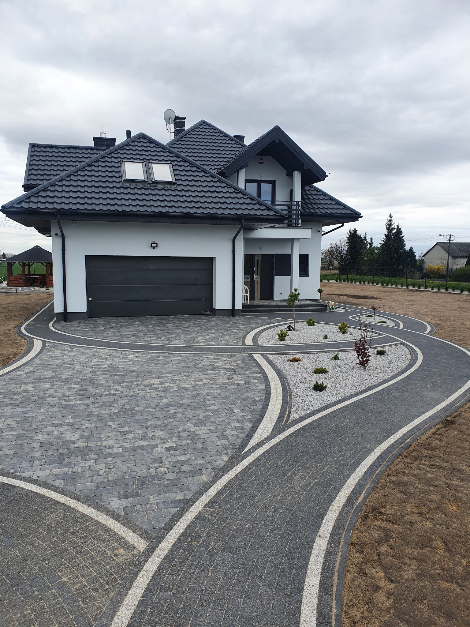 Nowoczesny dom z eleganckim podjazdem z kostki brukowej w odcieniach szarości, z białymi akcentami i rabatami z białym żwirem. Widok na budynek z garażem.