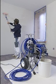 Malowanie ścian agregatem hydrodynamicznym: malarz w masce ochronnej, niebieski wąż, białe ściany, zabezpieczone okno, podłoga wyłożona folią ochronną.