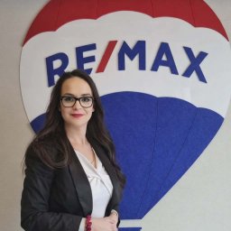EM Nieruchomości Ewa Mozdyniewicz - Kobieta w garniturze, z okularami, stoi na tle logo RE/MAX. Profesjonalny wizerunek agenta nieruchomości, budzący zaufanie. Jasne tło, dobre oświetlenie.