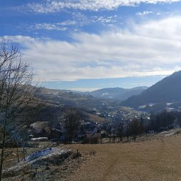 EM Nieruchomości Ewa Mozdyniewicz - Malowniczy krajobraz górski z widokiem na osadę, błękitne niebo z chmurami i drzewa na pierwszym planie. Zimowy klimat, naturalne światło.
