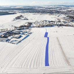EM Nieruchomości Ewa Mozdyniewicz - Zimowy widok z lotu ptaka na działkę o powierzchni 8033 mkw, z zaznaczonym na niebiesko przebiegiem drogi dojazdowej. W tle zabudowania i krajobraz.