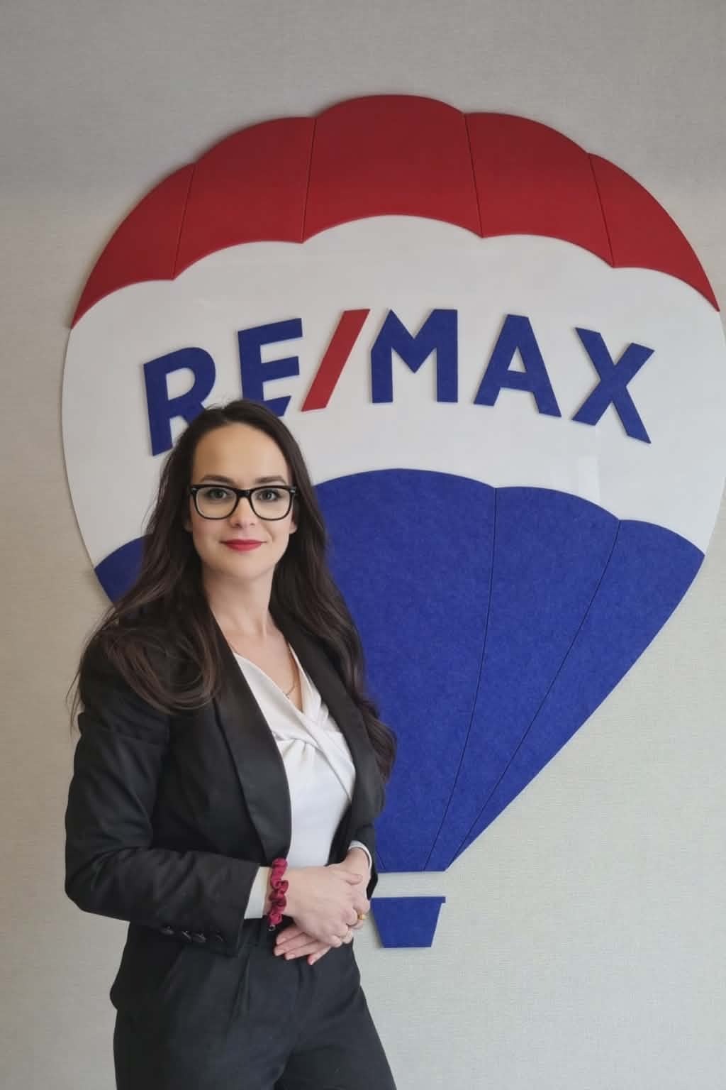 Kobieta w garniturze, z okularami, stoi na tle logo RE/MAX. Profesjonalny wizerunek agenta nieruchomości, budzący zaufanie. Jasne tło, dobre oświetlenie.