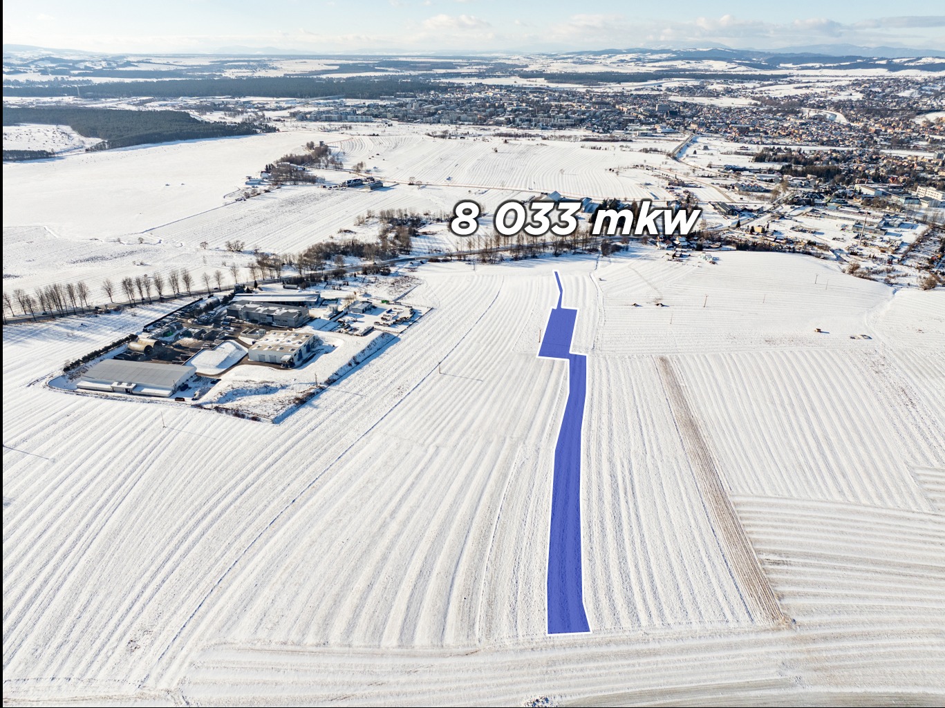 Zimowy widok z lotu ptaka na działkę o powierzchni 8033 mkw, z zaznaczonym na niebiesko przebiegiem drogi dojazdowej. W tle zabudowania i krajobraz.