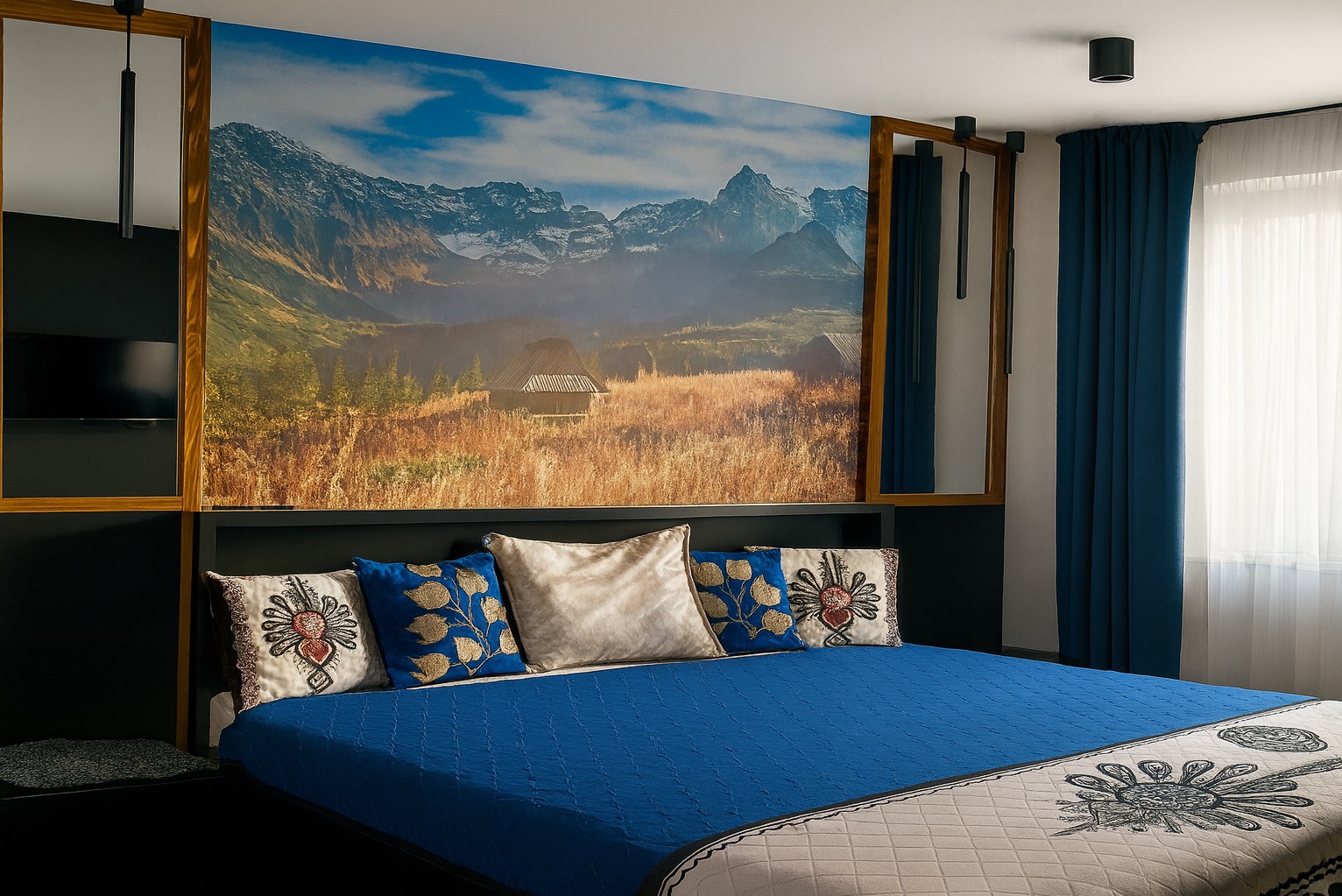 Sypialnia z fototapetą górskiego krajobrazu, niebieską narzutą i dekoracyjnymi poduszkami. Nowoczesne oświetlenie i drewniane ramy dodają wnętrzu charakteru.