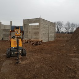 Siatrixstal Izabela Goździejewska - Żółta wiertnica hydrauliczna na gąsienicach przed wznoszonym ogrodzeniem z prefabrykatów betonowych o fakturze kamienia. W tle drzewa i hałda ziemi.