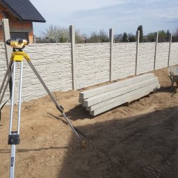 Siatrixstal Izabela Goździejewska - Montaż ogrodzenia betonowego z użyciem niwelatora. Widoczne elementy ogrodzenia, taczka i pracownik w tle. Etap budowy ogrodzenia z fakturą kamienia.