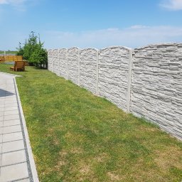 Siatrixstal Izabela Goździejewska - Długi, szary mur z betonu imitującego kamień, oddzielający trawnik od dalszej części ogrodu z drewnianymi meblami. Widok w słoneczny dzień.