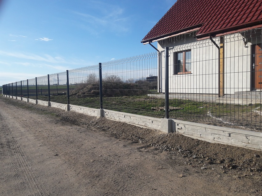 Długie, czarne ogrodzenie panelowe z podmurówką z elementami dekoracyjnymi, otaczające posesję z domem o czerwonym dachu, widziane z polnej drogi w słoneczny dzień.