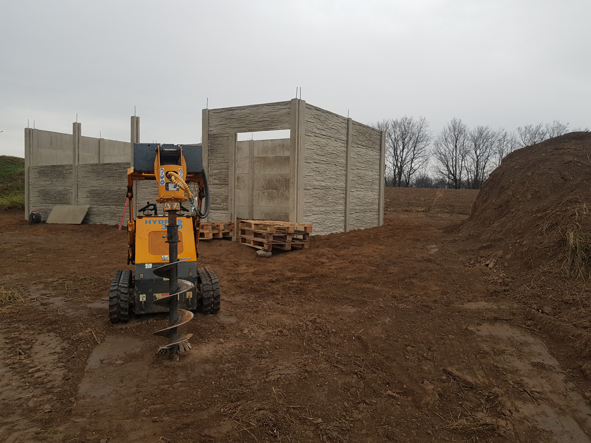 Żółta wiertnica hydrauliczna na gąsienicach przed wznoszonym ogrodzeniem z prefabrykatów betonowych o fakturze kamienia. W tle drzewa i hałda ziemi.