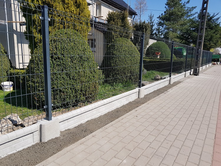 Nowoczesne ogrodzenie panelowe z podmurówką z cegły klinkierowej, szare słupki, widok z boku, kostka brukowa, zieleń w tle, słoneczny dzień.