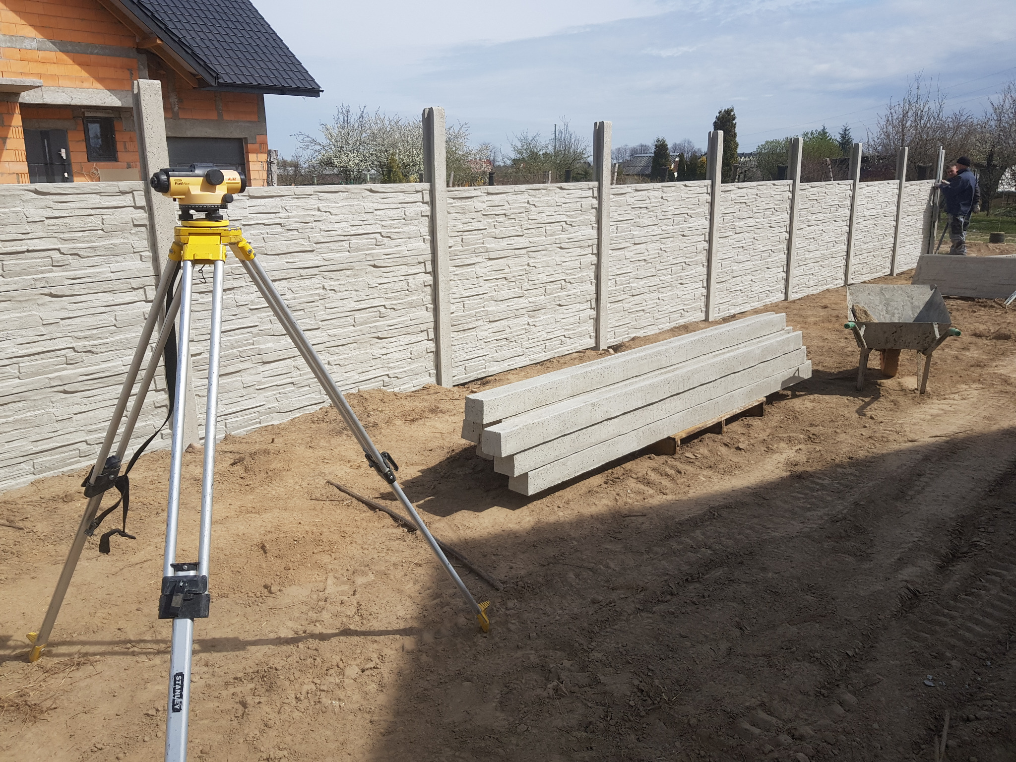 Montaż ogrodzenia betonowego z użyciem niwelatora. Widoczne elementy ogrodzenia, taczka i pracownik w tle. Etap budowy ogrodzenia z fakturą kamienia.
