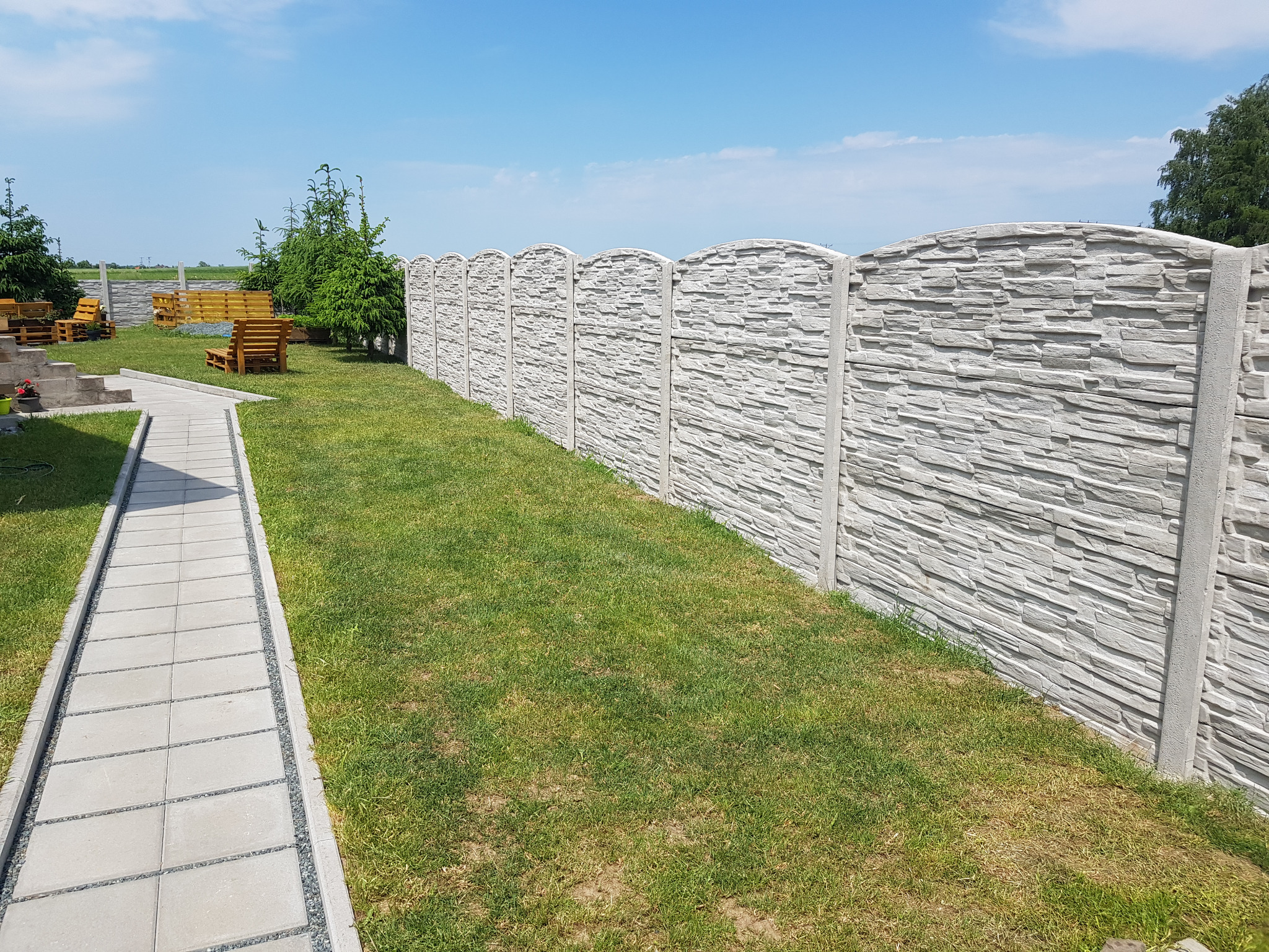 Długi, szary mur z betonu imitującego kamień, oddzielający trawnik od dalszej części ogrodu z drewnianymi meblami. Widok w słoneczny dzień.