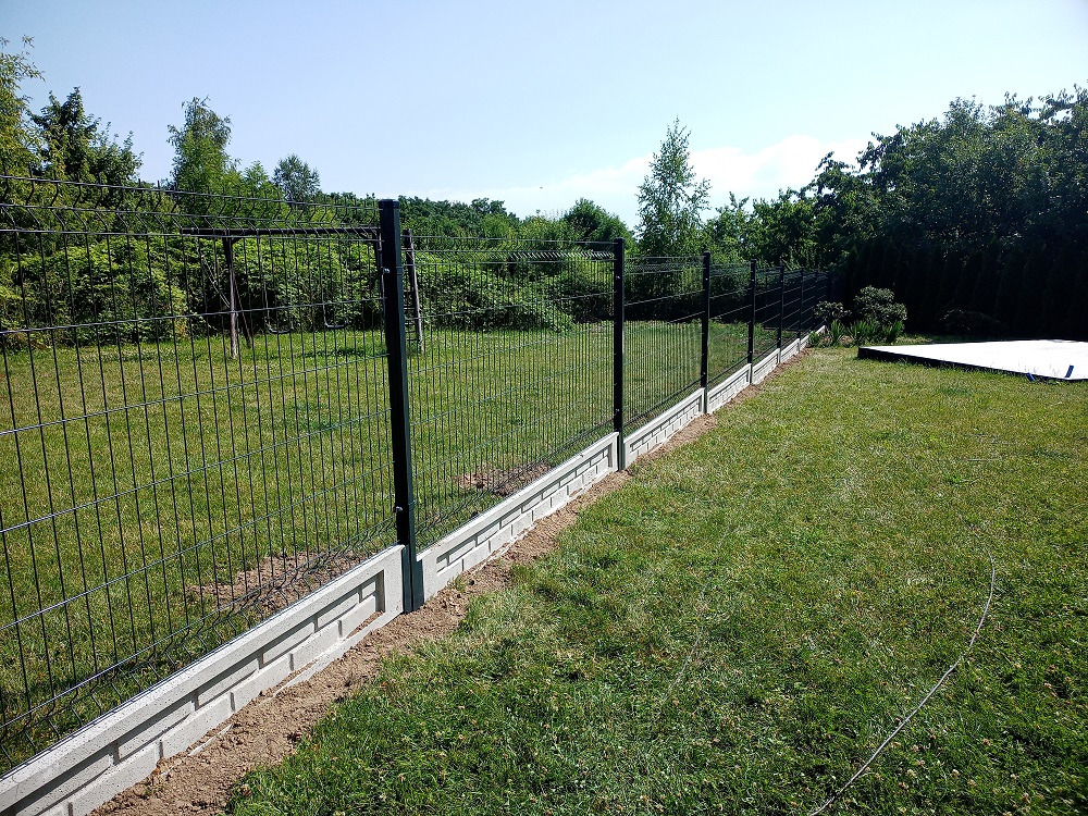 Długi, prosty odcinek ogrodzenia panelowego z podmurówką z szarej cegły, na tle zielonego trawnika i drzew. Widok z perspektywy lekko z dołu, w słoneczny dzień.