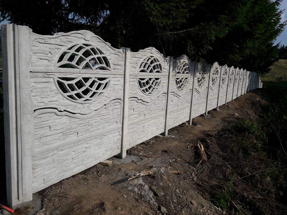 Długi, dekoracyjny, szary mur z prefabrykatów betonowych z okrągłymi ażurowymi zdobieniami, oddzielający teren. Widok z dołu, w tle zieleń drzew.