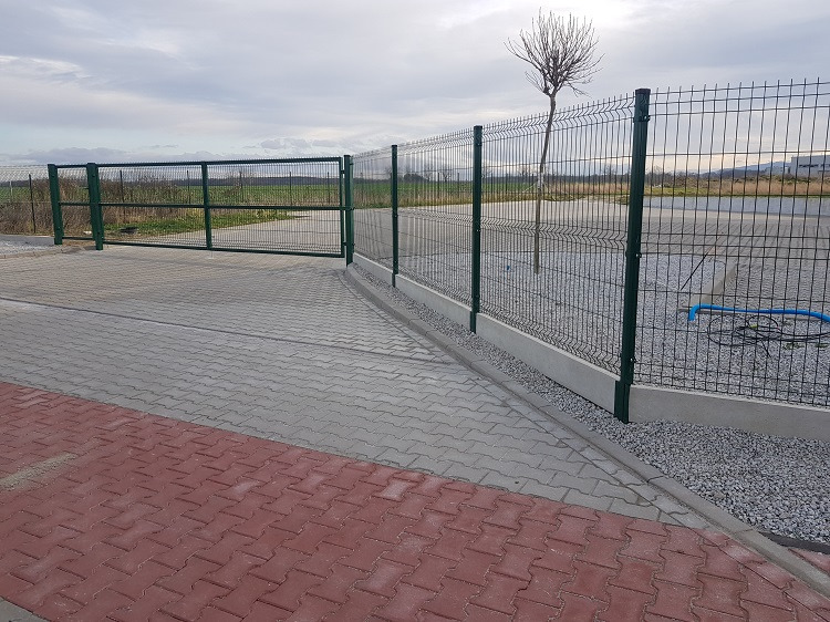 Nowoczesne ogrodzenie panelowe z bramą wjazdową, zielone słupki, podmurówka betonowa, kostka brukowa w dwóch kolorach. Widok na posesję i drzewo bez liści w tle.