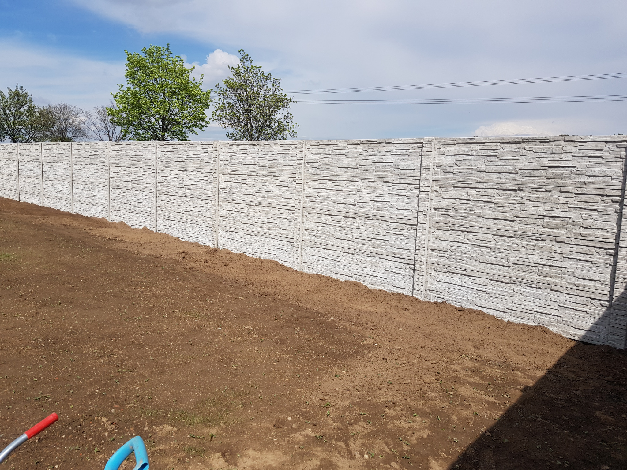 Długi, jasny, betonowy płot o fakturze kamienia, oddzielający teren z ziemią od zieleni drzew na tle błękitnego nieba z chmurami. Widoczne słupy i panele.