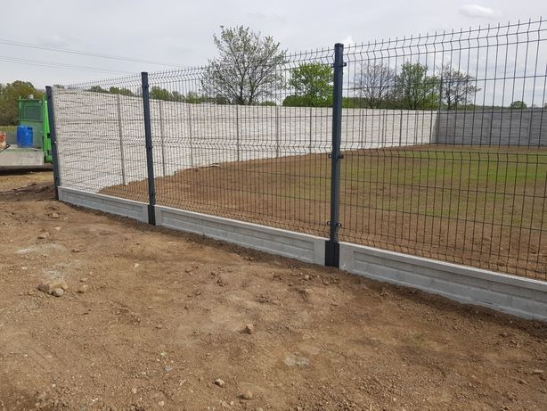 Panelowe ogrodzenie z podmurówką betonową na trawiastym terenie. Widoczne słupki i siatka ogrodzeniowa. W tle drzewa i błękitne niebo.