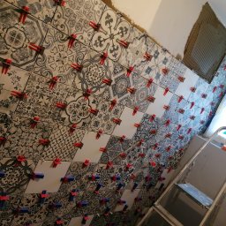 Artur Kamiński - Ściana w trakcie układania płytek z wzorami, z użyciem czerwonych i niebieskich klipsów poziomujących. Widoczny fragment ściany z nałożonym klejem i drabina.