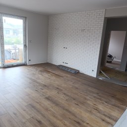 Artur Kamiński - Jasne panele podłogowe ułożone w nowym wnętrzu. Ściana z białej cegły dekoracyjnej z widocznymi puszkami elektrycznymi. Widok na sąsiednie pomieszczenie.