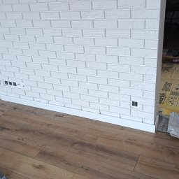 Artur Kamiński - Biała ściana z cegły dekoracyjnej, brązowa podłoga z paneli, widoczne puszki elektryczne i listwa przypodłogowa. W tle narzędzia na podłodze zabezpieczonej folią.