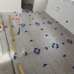 Artur Kamiński - Układanie beżowych płytek podłogowych z użyciem niebieskich klipsów poziomujących. Widoczna poziomica i rurka wystająca z podłogi. Prace w trakcie realizacji.