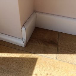 Artur Kamiński - Białe listwy przypodłogowe zamontowane w rogu pomieszczenia, na drewnopodobnej podłodze. Widoczne połączenie listew i fragment ściany w jasnym odcieniu.