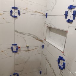 Artur Kamiński - Białe płytki imitujące marmur z niebieskimi klipsami poziomującymi, widoczna wnęka w ścianie i fragment poziomicy. Etap prac wykończeniowych w łazience.