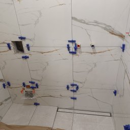 Artur Kamiński - Układanie płytek imitujących marmur w łazience, z użyciem klipsów poziomujących. Widoczny odpływ liniowy i narzędzia glazurnicze.