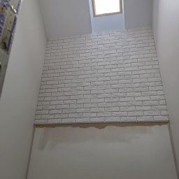 Artur Kamiński - Montaż białej cegły dekoracyjnej na ścianie pod oknem dachowym. Widoczny fragment drabiny. Prace wykończeniowe w jasnym pomieszczeniu.