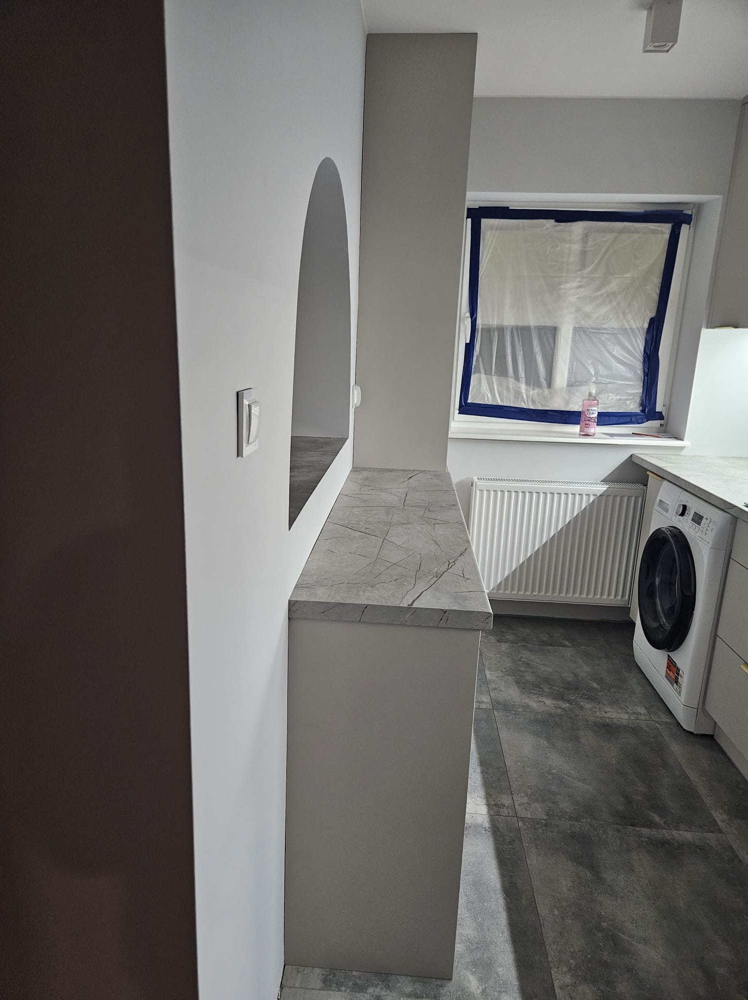 Jasna kuchnia z blatem imitującym marmur, okno zabezpieczone folią malarską, szare płytki podłogowe. Widoczna pralka i grzejnik.