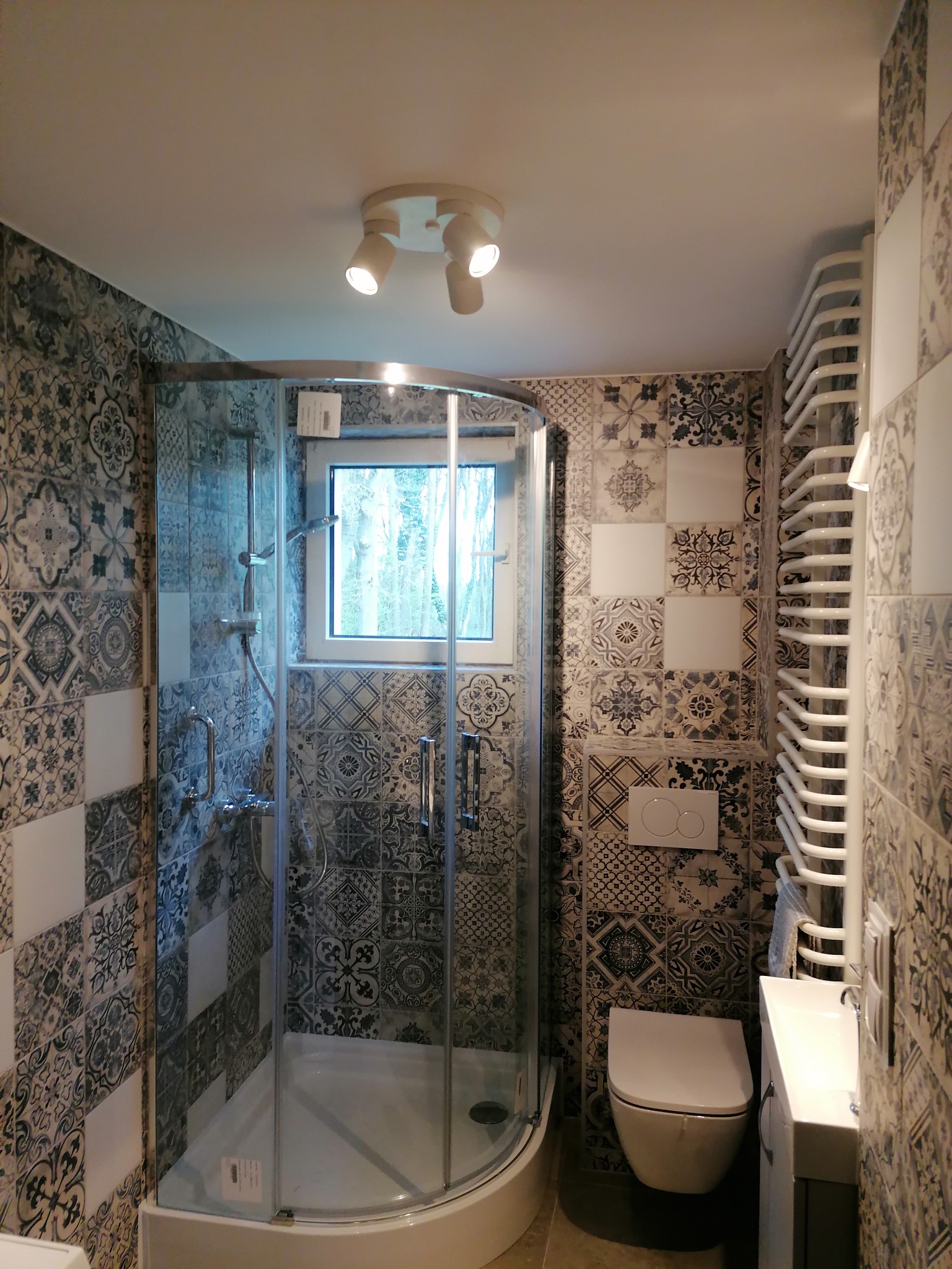 Wykończona łazienka z kabiną prysznicową, toaletą i grzejnikiem drabinkowym. Ściany wyłożone dekoracyjnymi płytkami w odcieniach szarości i bieli. Nowoczesne oświetlenie.