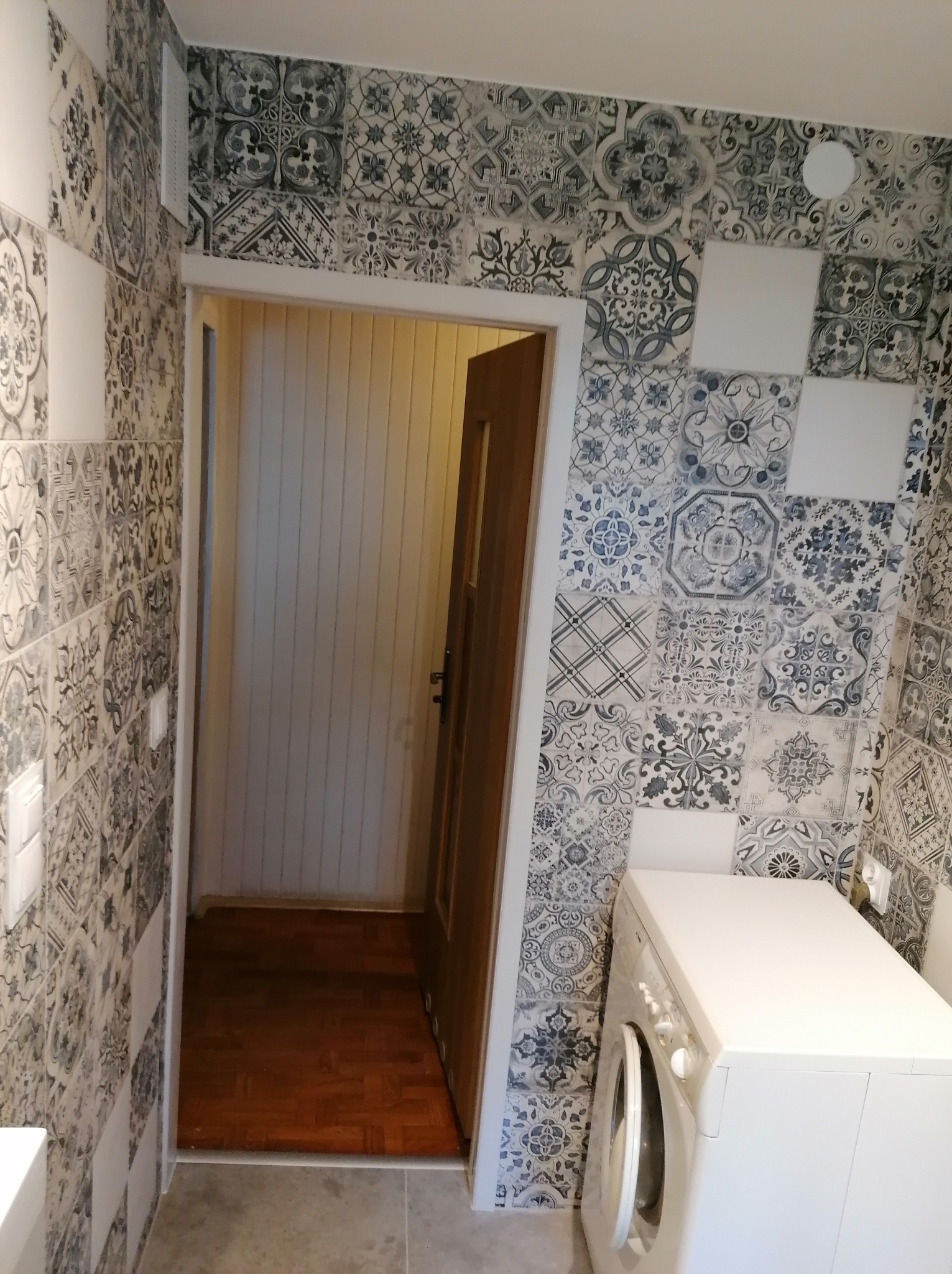 Wzorzyste płytki w odcieniach szarości i bieli zdobią ściany łazienki. Widoczna pralka i otwarte drzwi z drewnianą ościeżnicą. Podłoga wyłożona panelami.