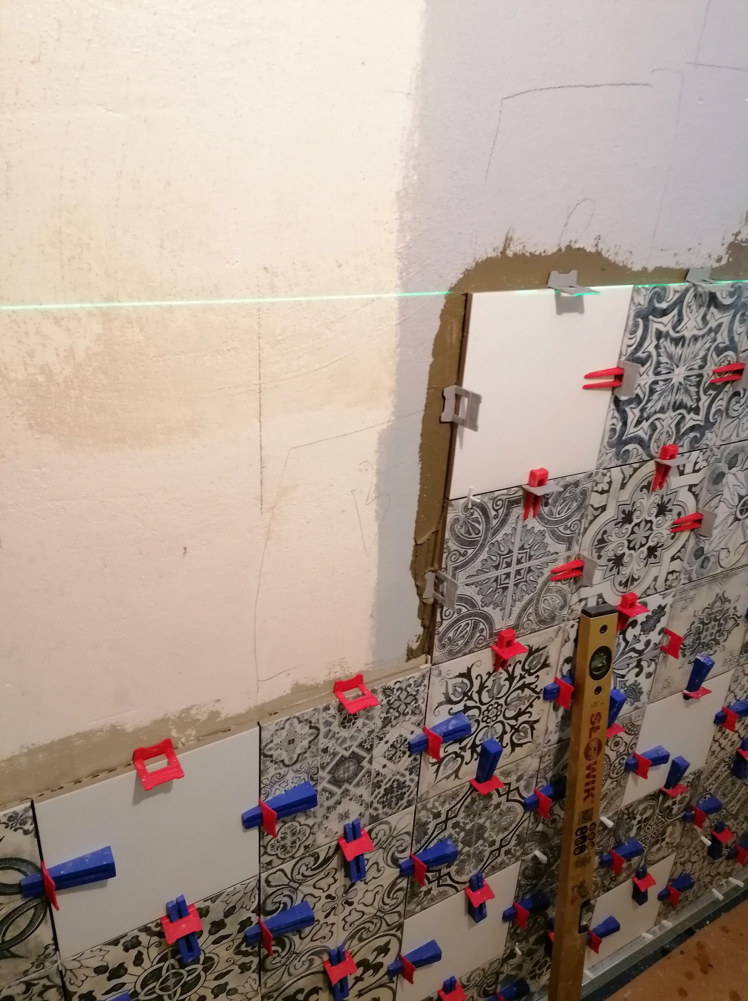 Układanie dekoracyjnych płytek na ścianie z użyciem poziomicy laserowej i klipsów montażowych. Widoczny fragment surowej ściany i zaprawa.