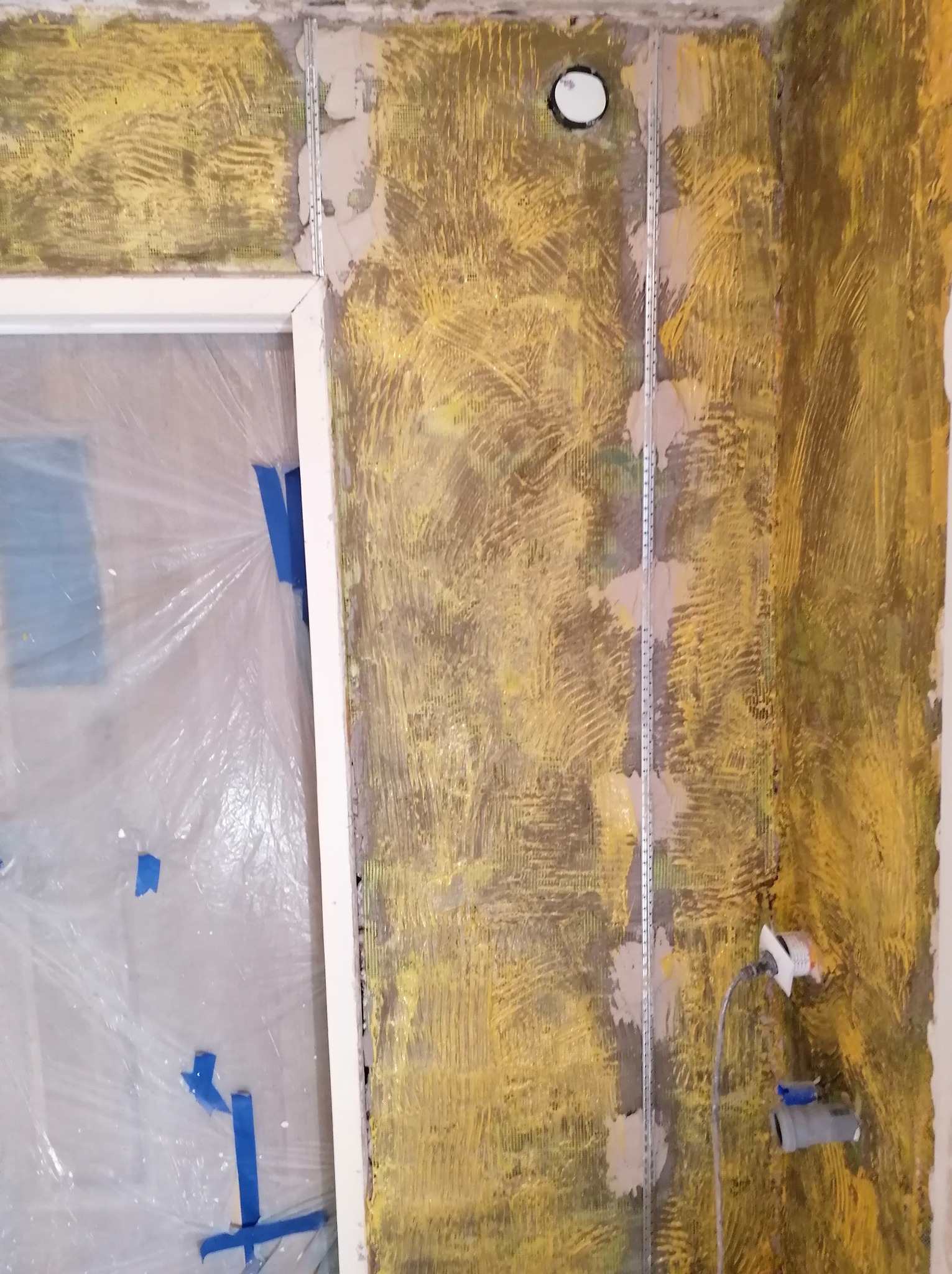 Żółta siatka na ścianie z profilami i otworem na instalację, zabezpieczona folią okienna. Etap przygotowania podłoża pod wykończenie.