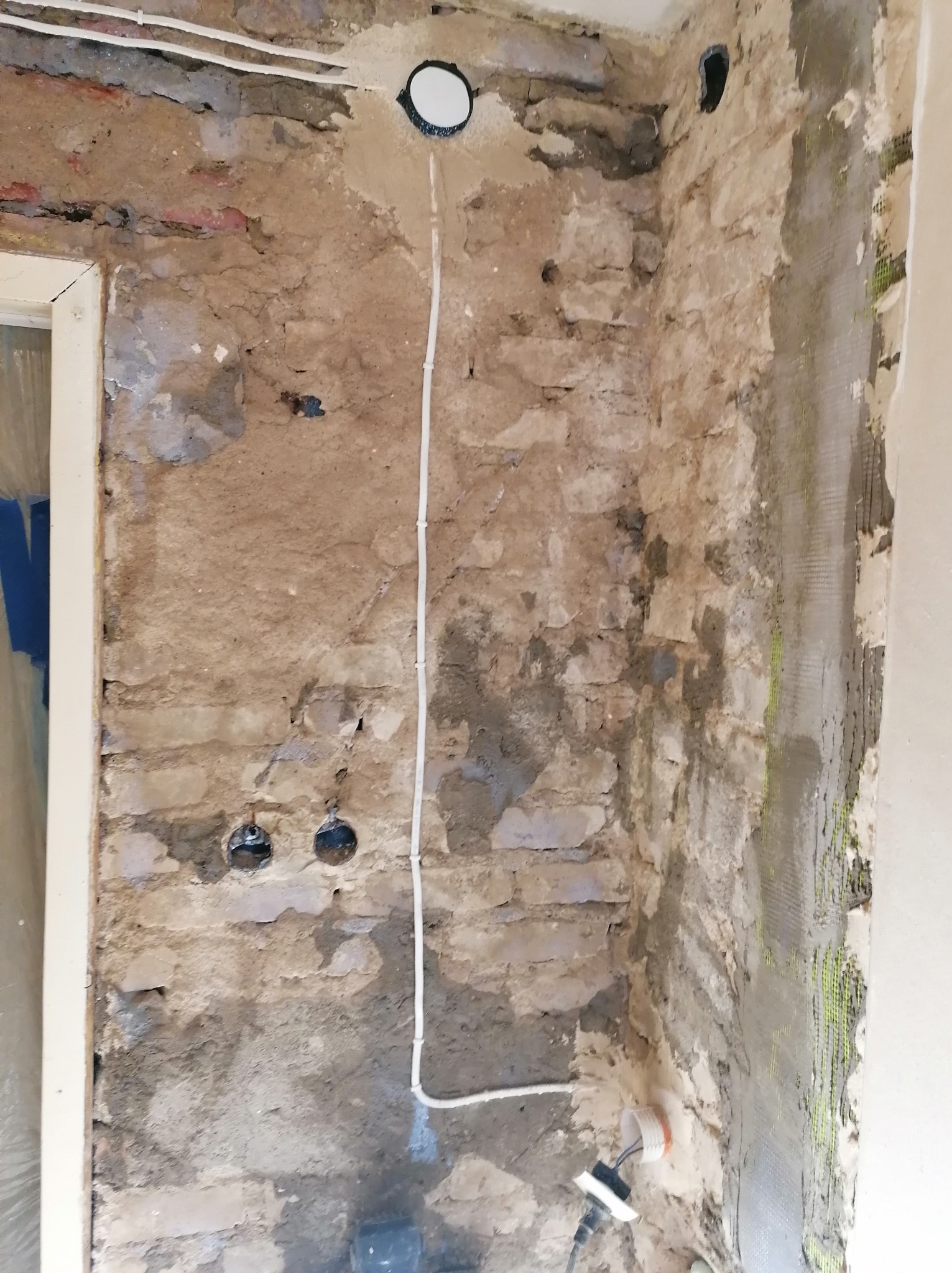 Odsłonięta ceglana ściana w trakcie prac przygotowawczych: instalacja elektryczna, siatka pod tynk na rogu. Widoczne otwory pod gniazdka i przewody.