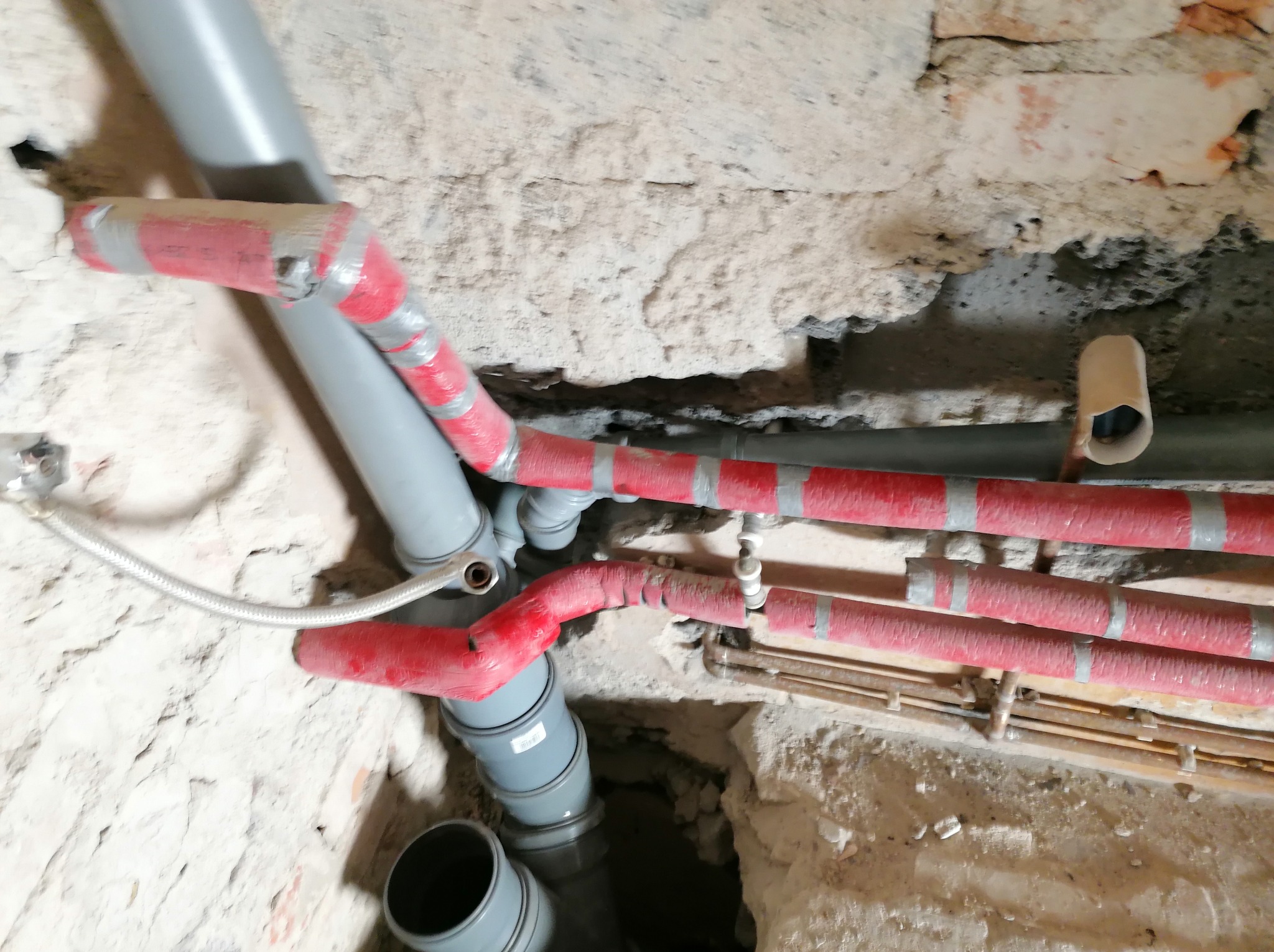 Szare rury kanalizacyjne i czerwone rury grzewcze, zabezpieczone taśmą, ułożone w bruzdach w surowej, ceglanej ścianie podczas remontu łazienki.