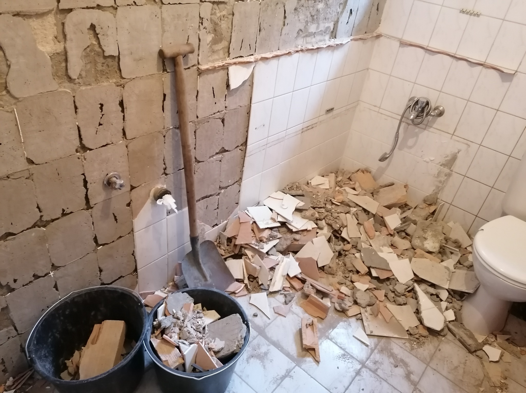 Demontaż płytek w łazience: ściany częściowo bez okładziny, gruz na podłodze, łopata oparta o ścianę, dwa wiadra z resztkami płytek, toaleta. Widok na remont w toku.