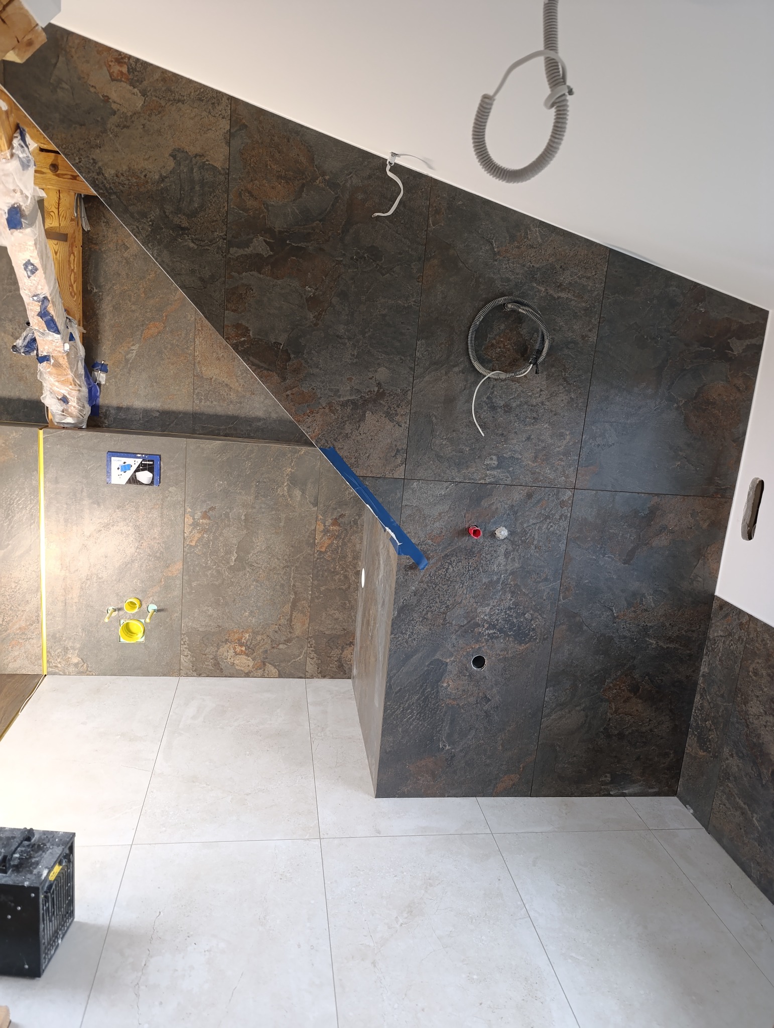 Ukończona łazienka na poddaszu z brązowo-szarymi płytkami na ścianach i jasnymi na podłodze. Widoczne instalacje i elementy montażowe. Ukośna ściana dodaje unikalnego charakteru.