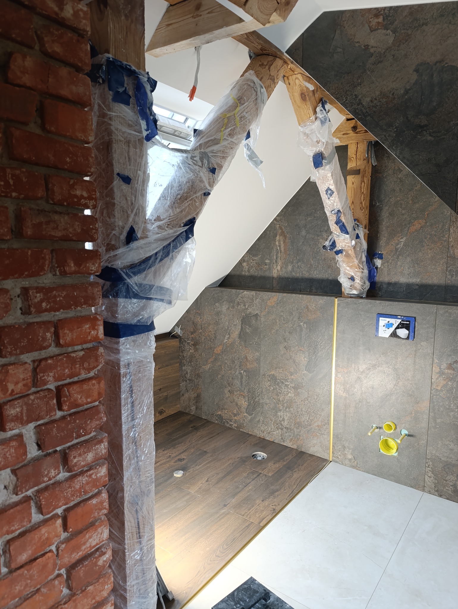 Wykończona łazienka w stylu rustykalnym z drewnianymi belkami zabezpieczonymi folią, brązowymi i białymi płytkami oraz elementami cegły. Nietypowe połączenie materiałów.