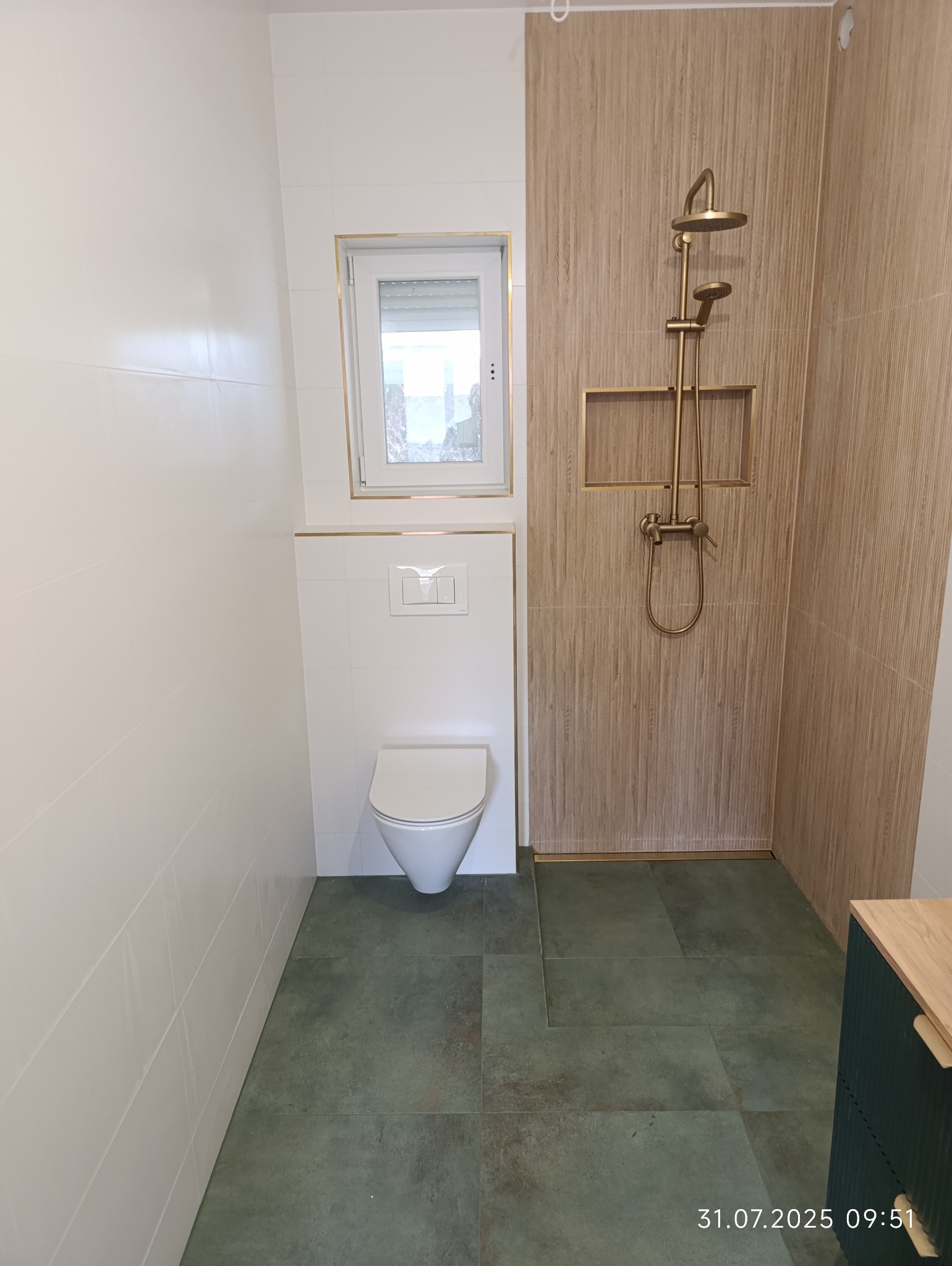 Nowoczesna łazienka po remoncie: białe płytki, drewnopodobne panele, złota armatura i zielona podłoga. Uwagę przykuwa minimalistyczna toaleta pod oknem.