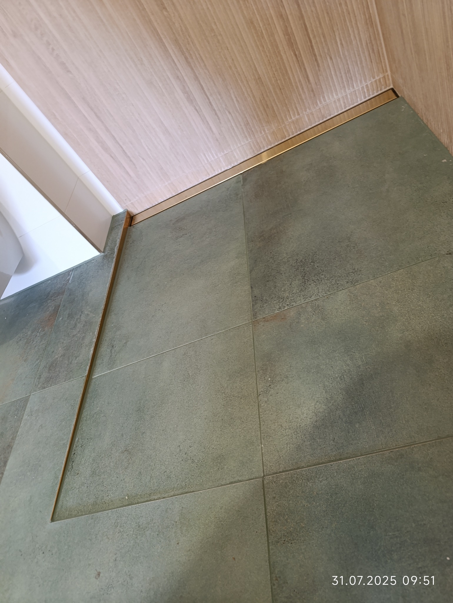 Wykończona łazienka z szarymi płytkami podłogowymi i drewnopodobnymi płytkami ściennymi, oddzielonymi złotą listwą. Widoczny fragment białej armatury.