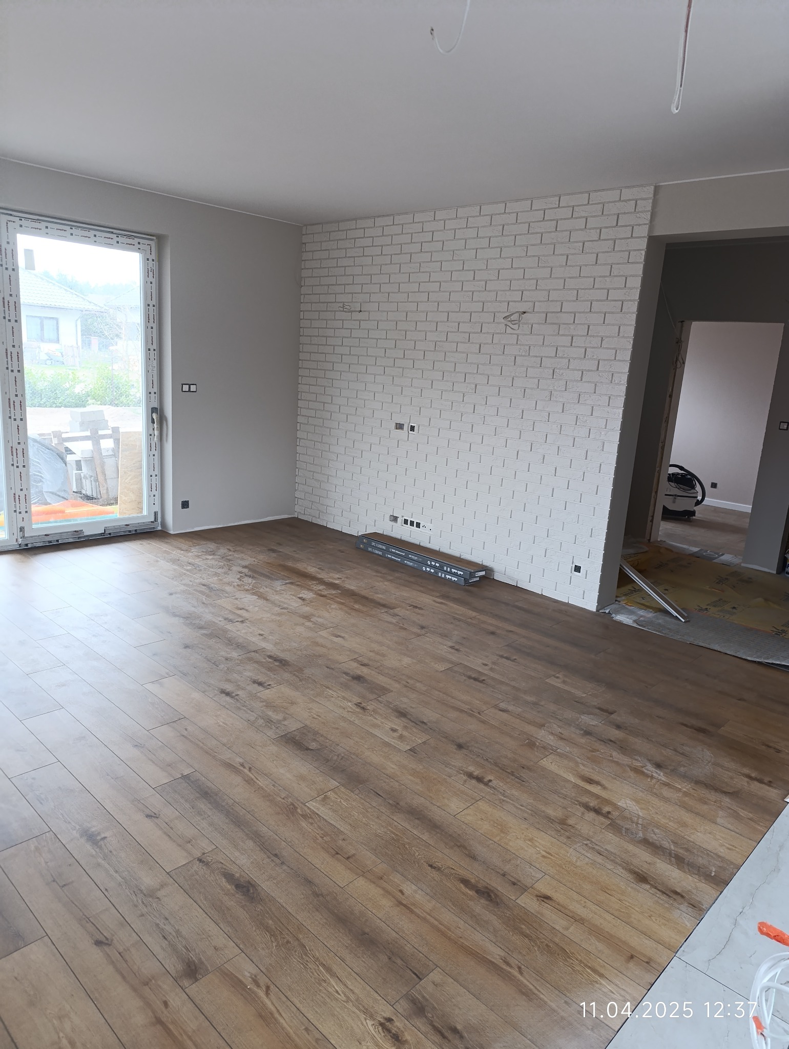 Jasne panele podłogowe ułożone w nowym wnętrzu. Ściana z białej cegły dekoracyjnej z widocznymi puszkami elektrycznymi. Widok na sąsiednie pomieszczenie.